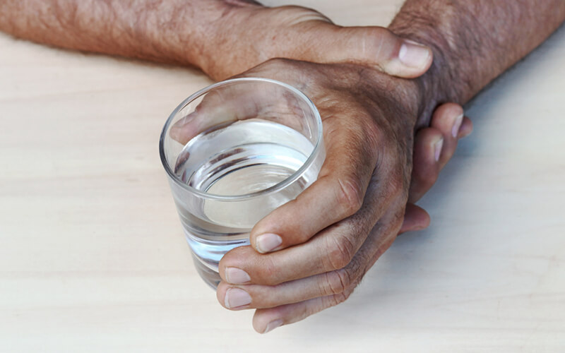 Closeup of elderly man’s hands struggling to hold a glass of water due to Parkinson’s disease